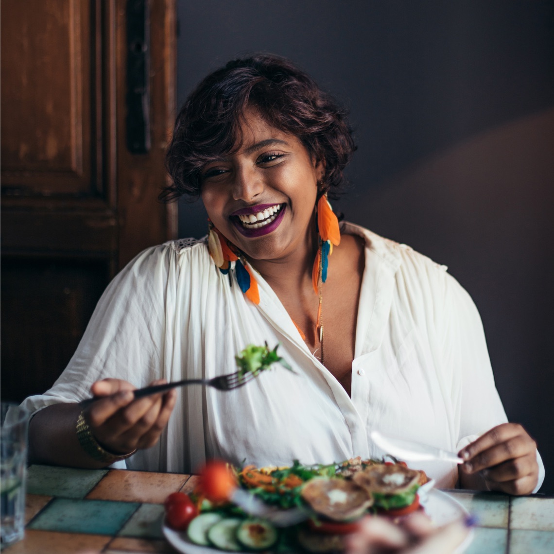 a woman wearing colorful earrings, smiling and eating a fresh salad